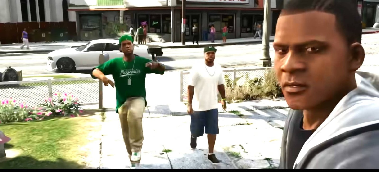Escena de un videojuego con tres personajes visibles en primer plano. El personaje a la izquierda lleva una camisa verde y una gorra verde, el personaje en el centro lleva una camisa blanca y pantalones cortos azules, y el personaje a la derecha lleva una sudadera gris. Al fondo, se ve un coche blanco, un edificio con un cartel y varios peatones en una calle urbana.